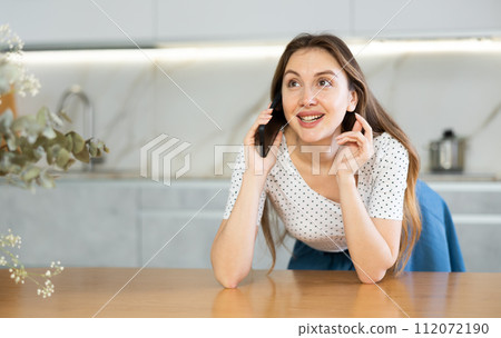 Woman with in comfortable clothing talking amiably on mobile phone while standing in home kitchen Woman with in comfortable clothing talking amiably on mobile phone while standing in home kitchen 112072190