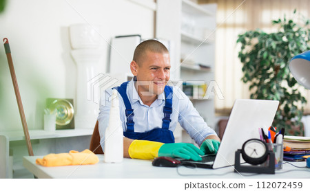 Office cleaner in overalls plays on laptop instead of cleaning office 112072459