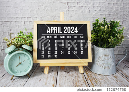 April 2024 monthly calendar and alarm clock on wooden background 112074768
