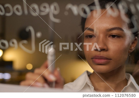 Closeup of young biracial female programmer writing code on glass wall in office late in evening 112076580