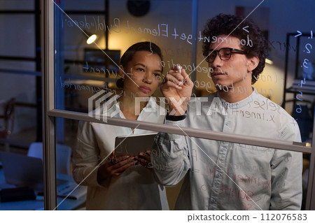 Young man and woman working together on code writing it on glass wall in IT company office late in evening 112076583