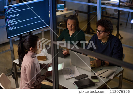 High angle view of ethnically diverse engineers working together on software in IT company office late in evening 112076596
