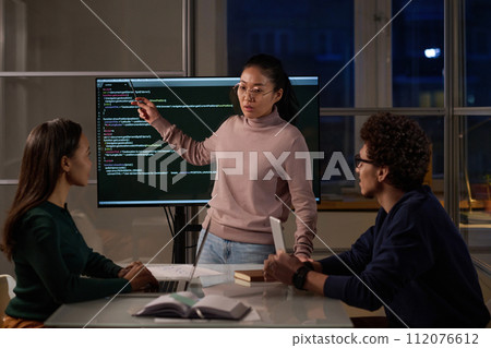Asian female specialist demonstrating code to her colleagues during meeting in IT company office late in evening Asian female specialist demonstrating code to her colleagues during meeting in IT company office late in evening 112076612