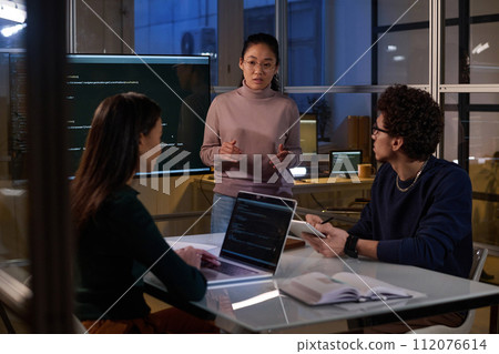 Asian programmer standing in front of colleagues discussing problems with computer code late in evening Asian programmer standing in front of colleagues discussing problems with computer code late in evening 112076614