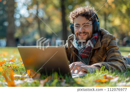 Smiling man wearing wireless headphones lying in grass with laptop in park 112076977