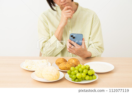 A middle-aged woman worries about food containing a lot of sugar (carbohydrates) and holding a smartphone. A middle-aged woman worries about food containing a lot of sugar (carbohydrates) and holding a smartphone. 112077119