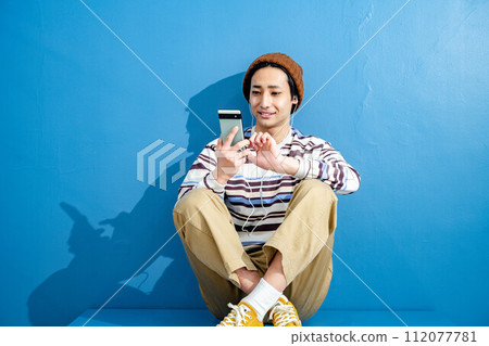 A young man wearing a hat and operating a smartphone against a blue background 112077781