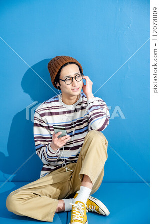 藍色背景下,戴著眼鏡和帽子玩智慧型手機的年輕人 藍色背景下,戴著眼鏡和帽子玩智慧型手機的年輕人 112078009