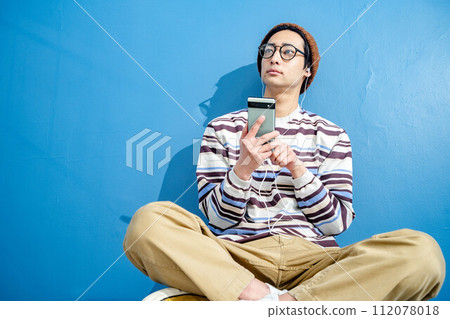 A young man playing with a smartphone while wearing glasses and a hat against a blue background A young man playing with a smartphone while wearing glasses and a hat against a blue background 112078018