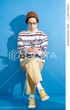 藍色背景下,戴著眼鏡和帽子玩智慧型手機的年輕人 藍色背景下,戴著眼鏡和帽子玩智慧型手機的年輕人 112078025