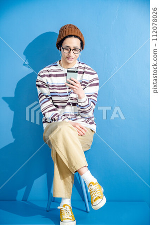 藍色背景下，戴著眼鏡和帽子玩智慧型手機的年輕人 112078026