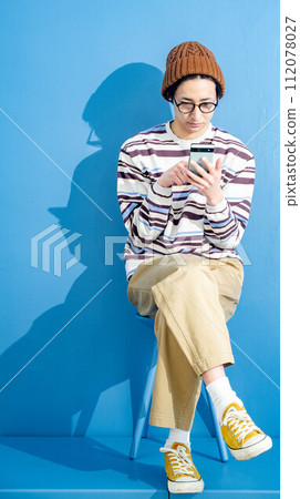 A young man playing with a smartphone while wearing glasses and a hat against a blue background 112078027