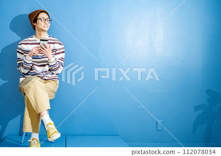 A young man playing with a smartphone while wearing glasses and a hat against a blue background 112078034