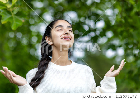 Close-up photo of a beautiful Indian woman standing in a park in a white sweater, raising her hands up and with her eyes closed, breathing and enjoying nature. 112079178