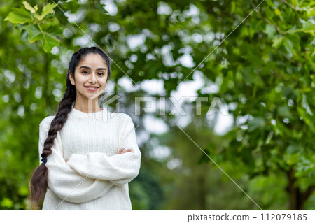 Close up portrait of a smiling Indian woman in a white sweater and braided hairstyle, standing in a park with her arms crossed on her chest and looking confidently at the camera. 112079185