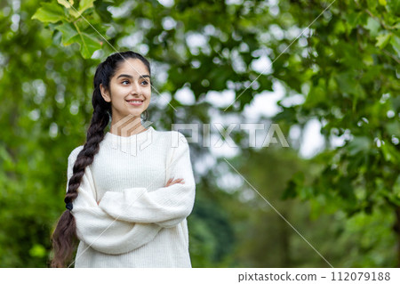 A happy and smiling Indian woman in a white sweater and a braided hairstyle stands in the park with her arms crossed, on her chest and looks confidently to the side. A happy and smiling Indian woman in a white sweater and a braided hairstyle stands in the park with her arms crossed, on her chest and looks confidently to the side. 112079188