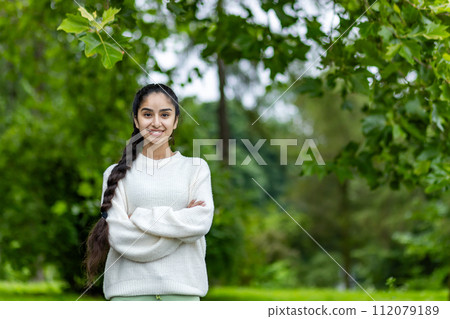 Portrait of a smiling Indian young girl in a white sweater doing sports and walking in the park. He stands with his hands crossed on his chest and looks confidently into the camera. Portrait of a smiling Indian young girl in a white sweater doing sports and walking in the park. He stands with his hands crossed on his chest and looks confidently into the camera. 112079189