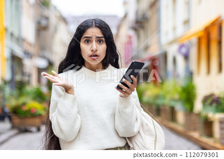 Portrait of an Indian serious and shocked woman standing in the city on the street, holding the phone in her hands and looking worriedly at the camera. Portrait of an Indian serious and shocked woman standing in the city on the street, holding the phone in her hands and looking worriedly at the camera. 112079301