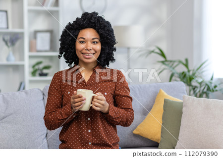 Close-up portrait of a smiling African American young woman relaxing on the couch at home, holding a cup of drink and looking at the camera. 112079390