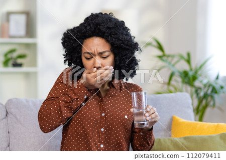 A sick young African-American woman sits at home on the sofa and drinks pills, holds a glass of water in her hands, feels bad. 112079411