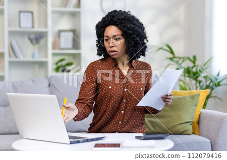 Upset African American woman sitting on sofa at home, holding documents in hands and looking worriedly at laptop screen. Checks accounts, works remotely. 112079416