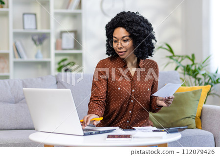 Concentrated and smiling young African American woman working remotely at home with family budget documents and laptop, checking household accounts and family budget. Concentrated and smiling young African American woman working remotely at home with family budget documents and laptop, checking household accounts and family budget. 112079426