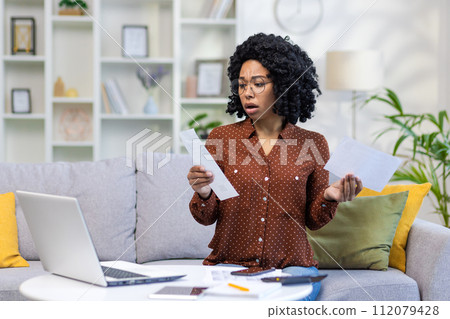 Upset and worried young African American woman sitting on sofa at home and looking at documents and financial accounts with family budget. He holds papers in his hands and works remotely on a laptop. 112079428