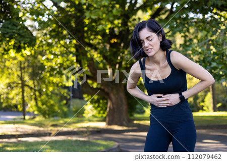 A young woman is standing in the park outdoors, bent over and holding her stomach, feeling severe pain after running and doing sports. 112079462