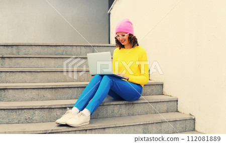 Portrait of stylish modern young woman working with laptop in colorful clothes in the city 112081889