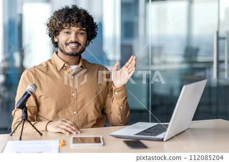 Confident islamic male sitting at workplace with laptop, tablet and professional microphone while smiling at camera. Productive blogger getting ready for recording new content in shared coworking. Confident islamic male sitting at workplace with laptop, tablet and professional microphone while smiling at camera. Productive blogger getting ready for recording new content in shared coworking. 112085204