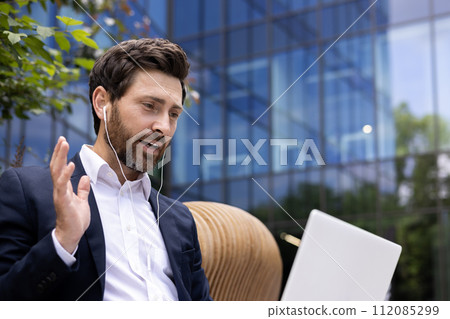 Confident man in suit engaging in video conference with colleagues using laptop and earphones outdoors. 112085299