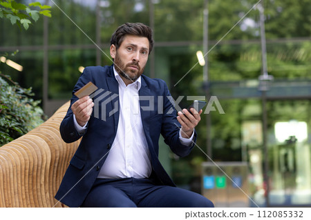 A businessman looks confused with a credit card and smartphone in hand, sitting outside an office, portraying finance issues. A businessman looks confused with a credit card and smartphone in hand, sitting outside an office, portraying finance issues. 112085332