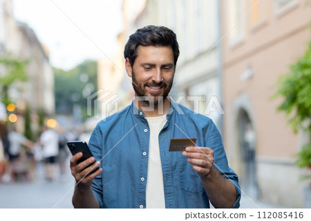 A smiling mature man with a beard uses a smartphone and credit card on a bustling city street as evening sets in. 112085416