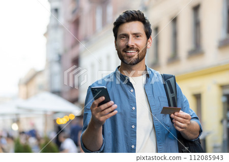 Portrait of a young male tourist standing on a city street with a backpack, holding a credit card and a mobile phone. Smiling and looking at the camera. 112085943