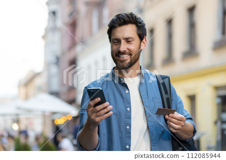 A traveling young man stands on a city street with a backpack, uses a credit card and a phone. Make online orders, reservations, shopping. A traveling young man stands on a city street with a backpack, uses a credit card and a phone. Make online orders, reservations, shopping. 112085944