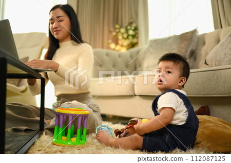 Cute little sitting on carpet near busy young beautiful mother working remotely on laptop Cute little sitting on carpet near busy young beautiful mother working remotely on laptop 112087125