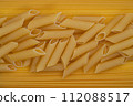 Scattered uncooked penne pasta lies on a yellow striped background, providing a textured look and culinary appeal. 112088517
