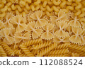 A colorful assortment of uncooked fusilli and farfalle pasta spread out, creating a vibrant yellow textured background. 112088524