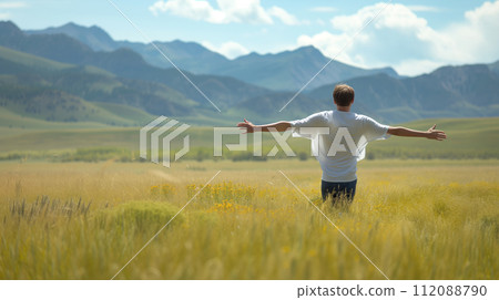 A man stands with open arms in a golden meadow, with the majestic mountains in the background under a clear sky. 112088790