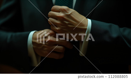 Detailed view of a man's hands as he carefully adjusts the cufflinks on his suit's sleeve, depicting sophistication and preparation. Detailed view of a man's hands as he carefully adjusts the cufflinks on his suit's sleeve, depicting sophistication and preparation. 112088805