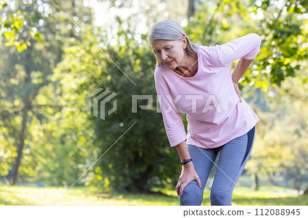 Active senior woman in sportswear touching her knee in discomfort while exercising in a sunlit park, depicting health issues. Active senior woman in sportswear touching her knee in discomfort while exercising in a sunlit park, depicting health issues. 112088945