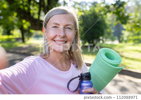 Healthy mature woman with a yoga mat and water bottle, smiling in a sunlit park. Fitness and wellbeing concept. 112088971