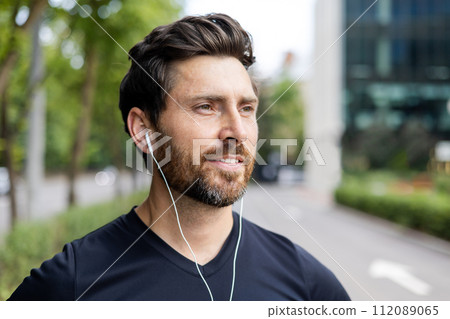 A smiling young man is standing on the street of the city wearing headphones and looking to the side. The athlete and instructor is doing sports and resting. 112089065