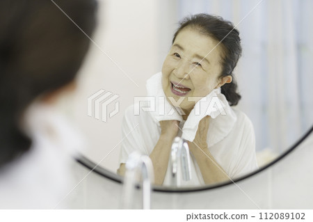 Smiling senior woman washing her face in the morning and wiping her face with a towel 112089102