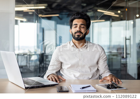 Relaxed young Indian man sitting in office at desk with laptop and documents and resting with closed eyes. Relaxed young Indian man sitting in office at desk with laptop and documents and resting with closed eyes. 112089122