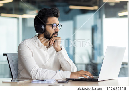Serious and focused Indian young man working in the office on a laptop. He is wearing headphones and is sitting at his desk. Serious and focused Indian young man working in the office on a laptop. He is wearing headphones and is sitting at his desk. 112089126