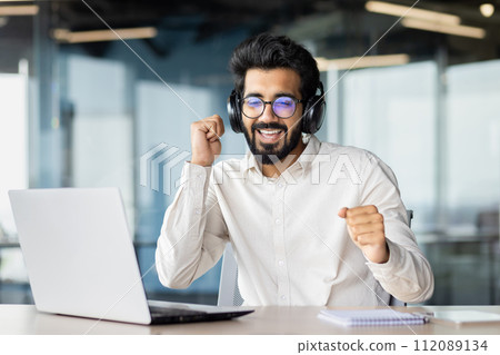 An office worker, a young Indian businessman sits in the office at the desk in headphones in front of the laptop, dances, moves his body and hands, relaxes. An office worker, a young Indian businessman sits in the office at the desk in headphones in front of the laptop, dances, moves his body and hands, relaxes. 112089134