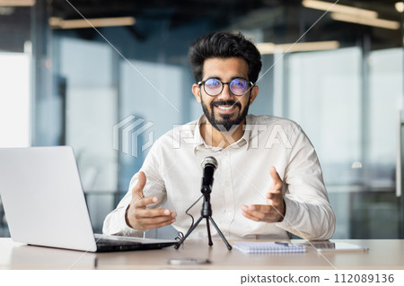 Smiling and successful young indian male businessman sitting in the office at the table in front of the microphone and having an online meeting, business training talking to the camera. Smiling and successful young indian male businessman sitting in the office at the table in front of the microphone and having an online meeting, business training talking to the camera. 112089136