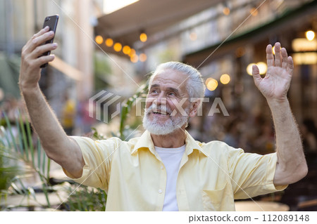 A cheerful elderly man in a yellow shirt waves while making a video call on his phone, enjoying the evening. 112089148