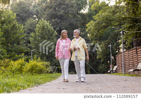 An elderly couple strolls hand in hand, exuding comfort and joy in a tranquil park, embodying lasting love and companionship. 112089157
